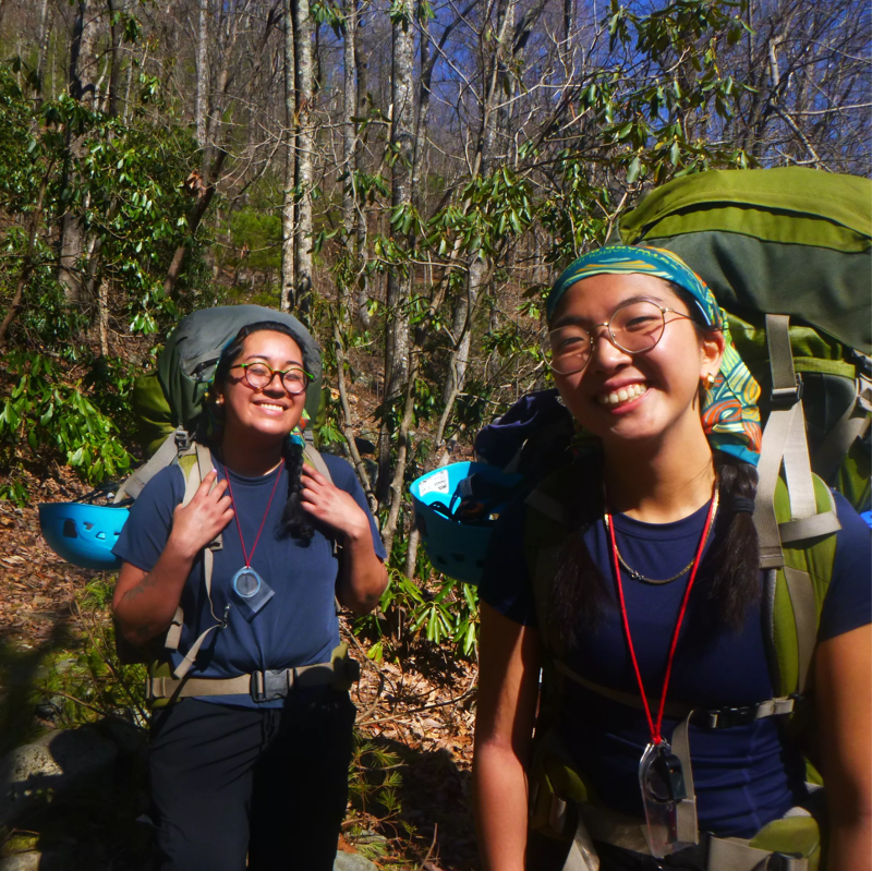 Two young women are hiking in a forest. They are wearing large backpacks and carrying gear, suggesting they are on a multi-day trip. Both women are smiling and appear to be enjoying their adventure. The forest is dense with trees, and the lighting indicates it is daytime.